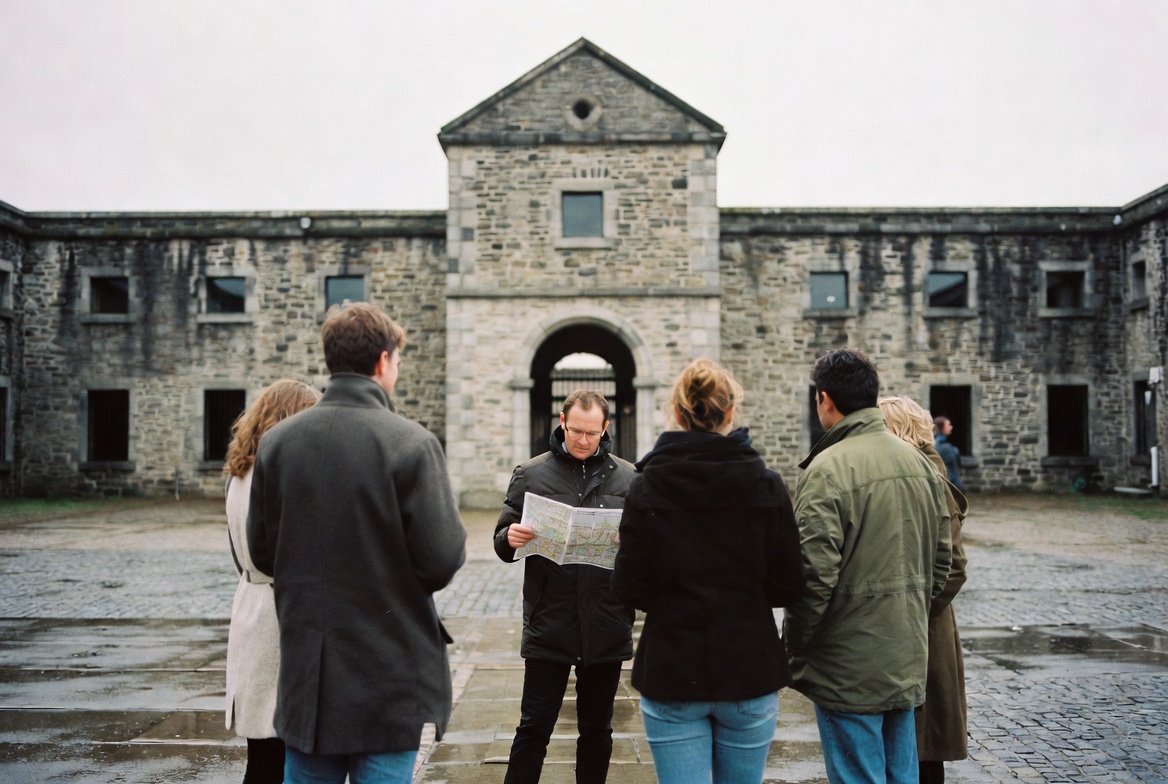 A walking group gathered on the cobbled forecourt of Kilmainham Gaol, listening to a guide point toward the upper windows.
